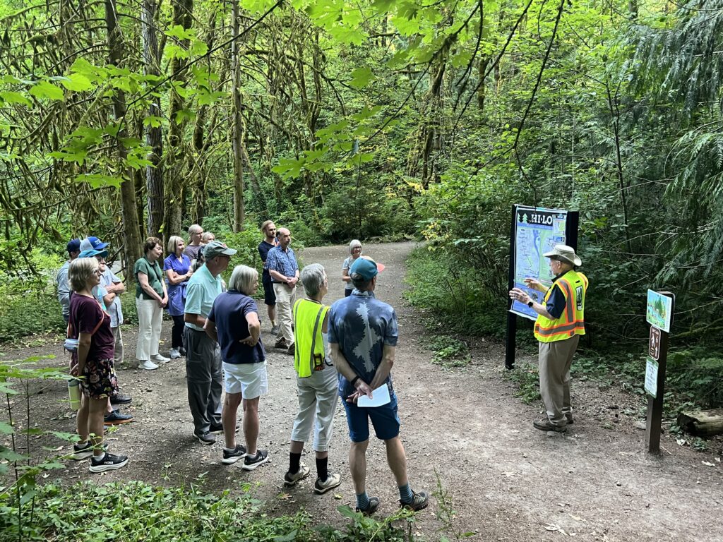 15 people standing in a forest listening to a man presenting a trail sign.